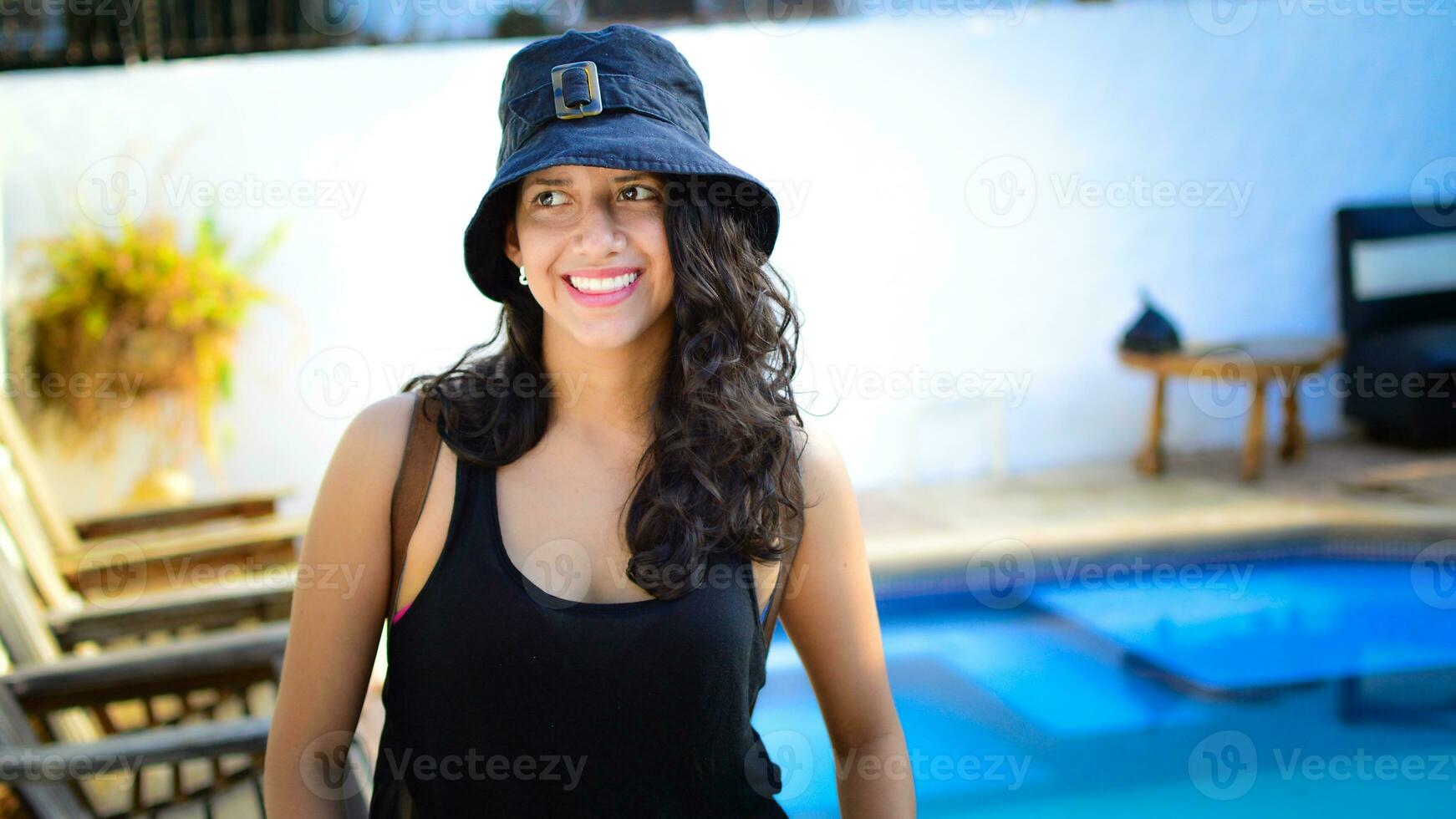 Pretty girl with hat smiling near a swimming pool, latin girl smiling by a swimming pool with muskoka chairs photo
