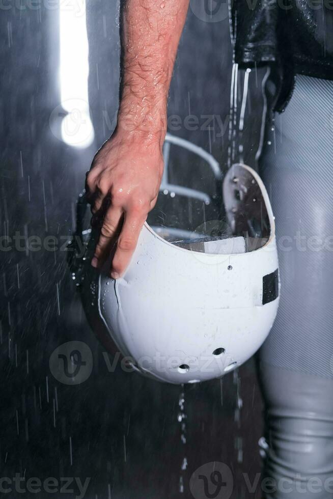 Close up of American Football Athlete Warrior Standing on a Field focus on his Helmet and Ready to Play. Player Preparing to Run, Attack and Score Touchdown. Rainy Night with Dramatic lens flare and rain drops. photo