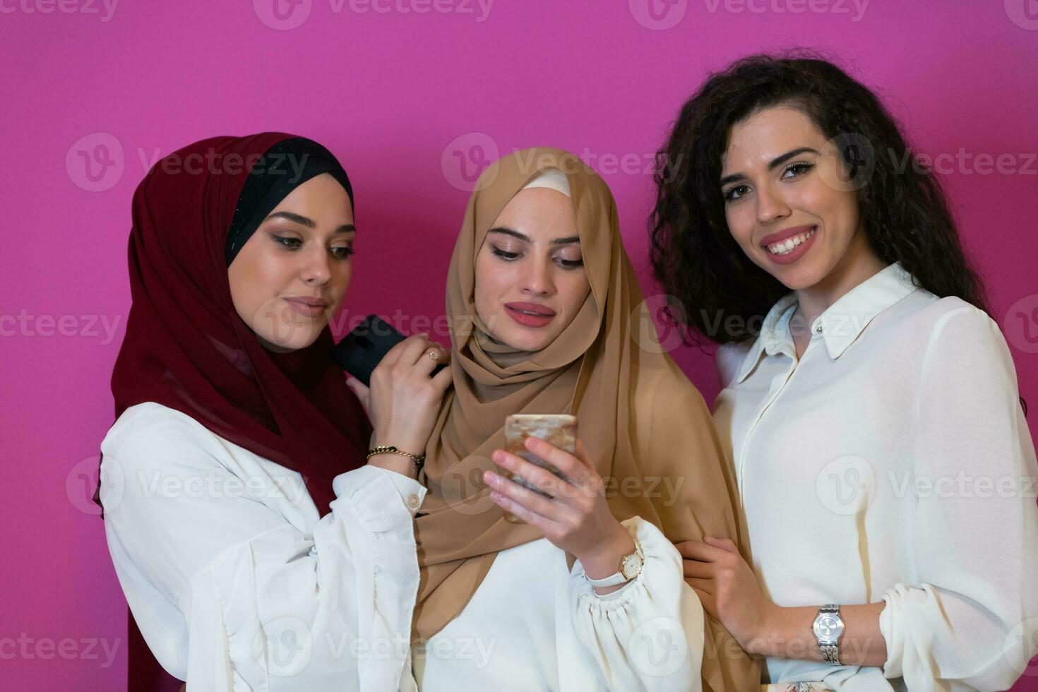 Group of beautiful muslim women two of them in fashionable dress with hijab using mobile phone isolated on pink background representing modern islam fashion technology and ramadan kareem concept photo