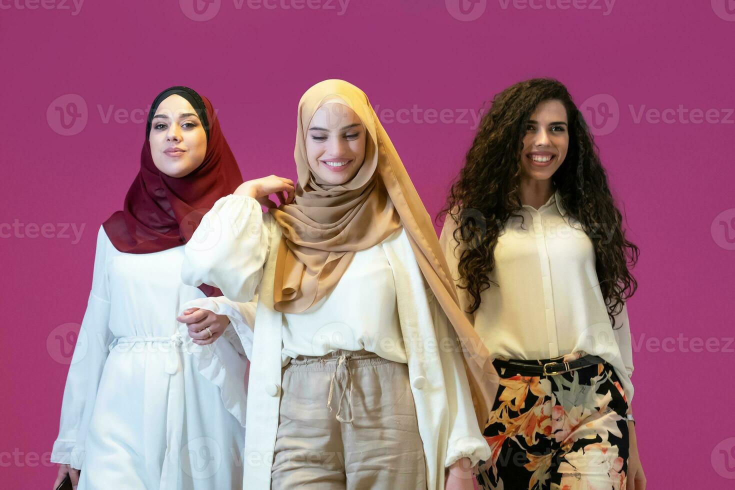Group portrait of beautiful muslim women two of them in fashionable dress with hijab isolated on pink background representing modern islam fashion and ramadan kareem concept photo