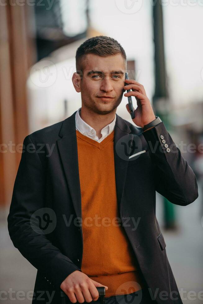 ir a la terminal del aeropuerto. viajero de negocios confiado caminando por las calles de la ciudad y tirando de su maleta bebiendo café y hablando en el teléfono inteligente foto