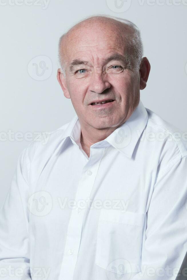 Portrait of an older man wearing a white shirt on a gray background. A healthy old man looks at a camera isolated over a gray wall. An older man smiles at the camera, a happy old man. photo