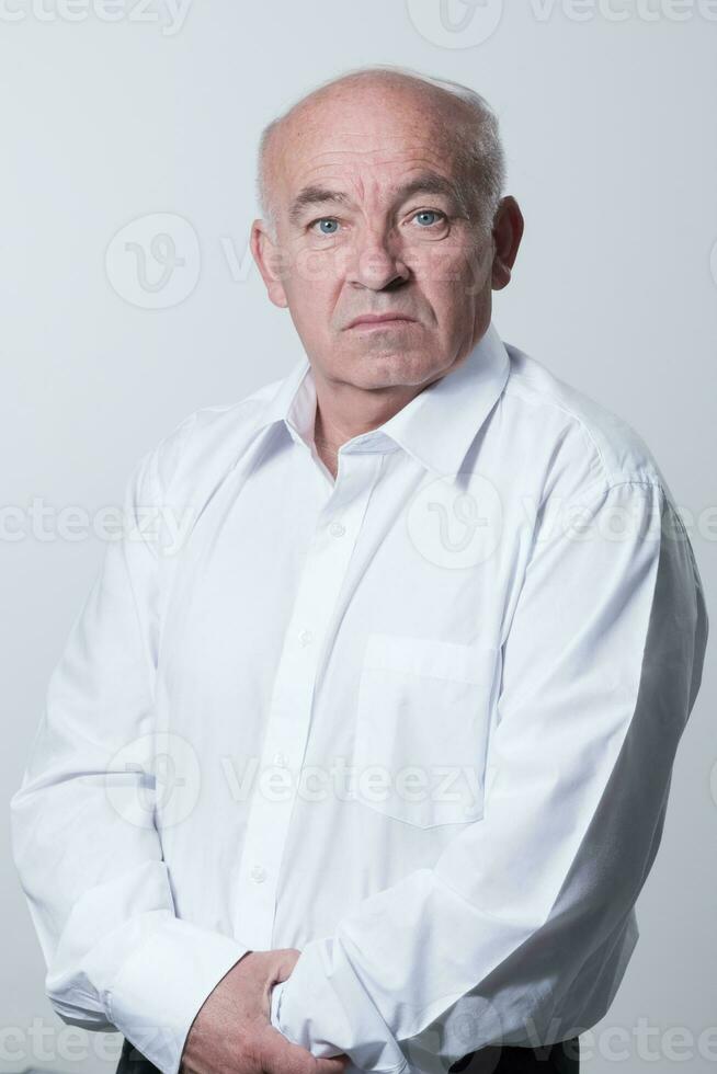 Portrait of an older man wearing a white shirt on a gray background. A healthy old man looks at a camera isolated over a gray wall. An older man smiles at the camera, a happy old man. photo