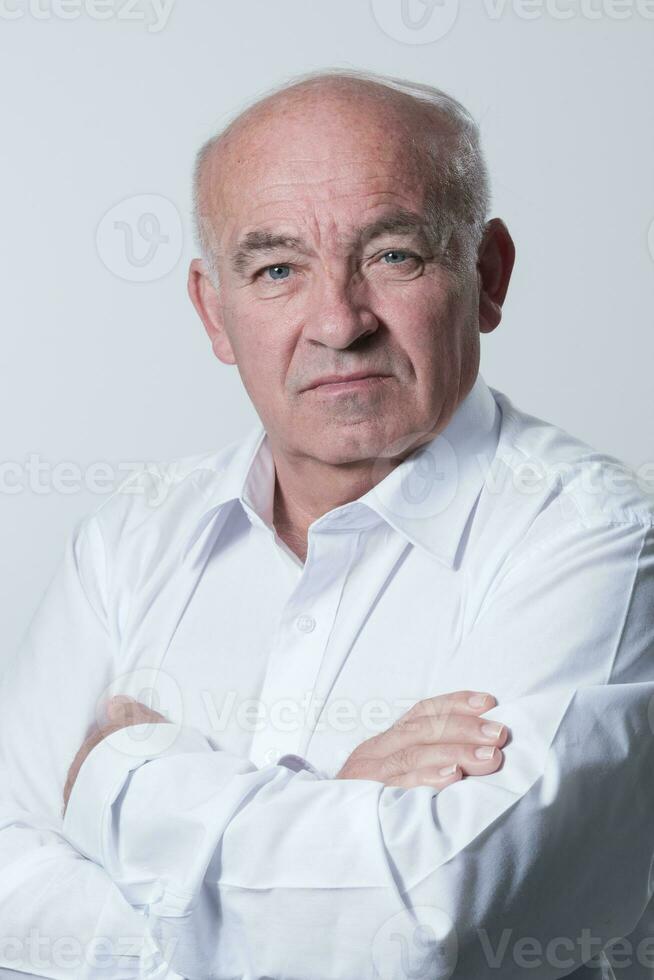 Confident senior man in white shirt crossing hands on chest and looking at camera while standing against gray background. Self confident senior isolated white studio shoot. photo