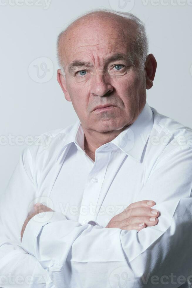 Confident senior man in white shirt crossing hands on chest and looking at camera while standing against gray background. Self confident senior isolated white studio shoot. photo