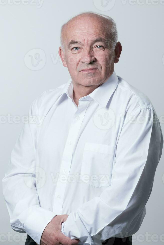 Portrait of an older man wearing a white shirt on a gray background. A healthy old man looks at a camera isolated over a gray wall. An older man smiles at the camera, a happy old man. photo