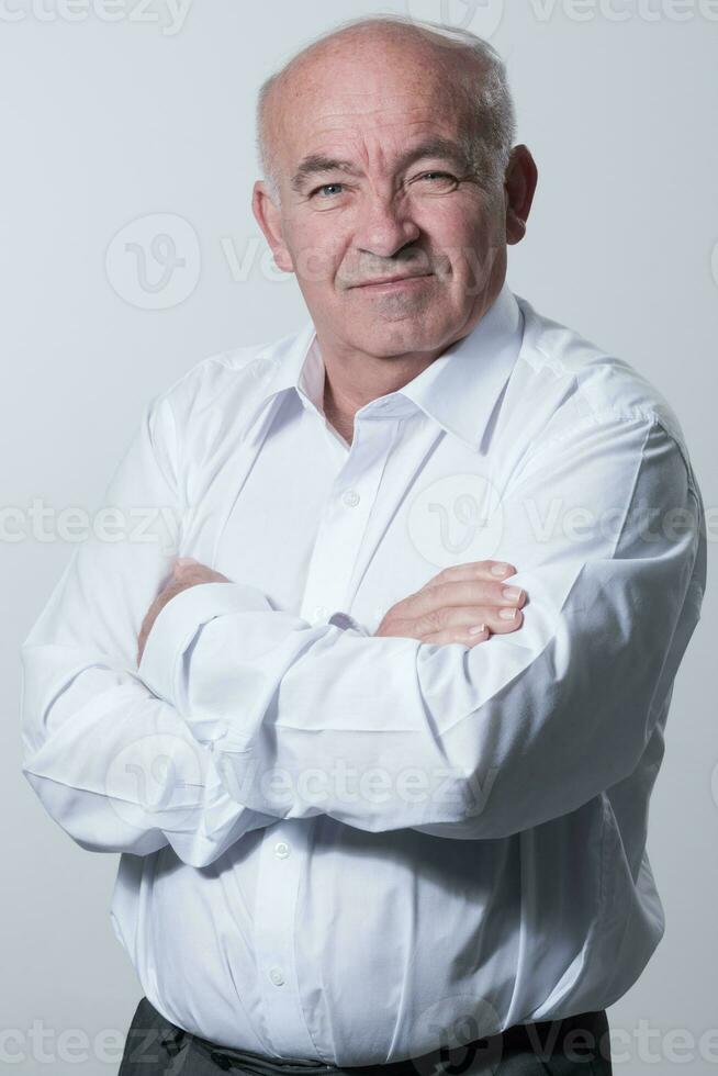 Confident senior man in white shirt crossing hands on chest and looking at camera while standing against gray background. Self confident senior isolated white studio shoot. photo