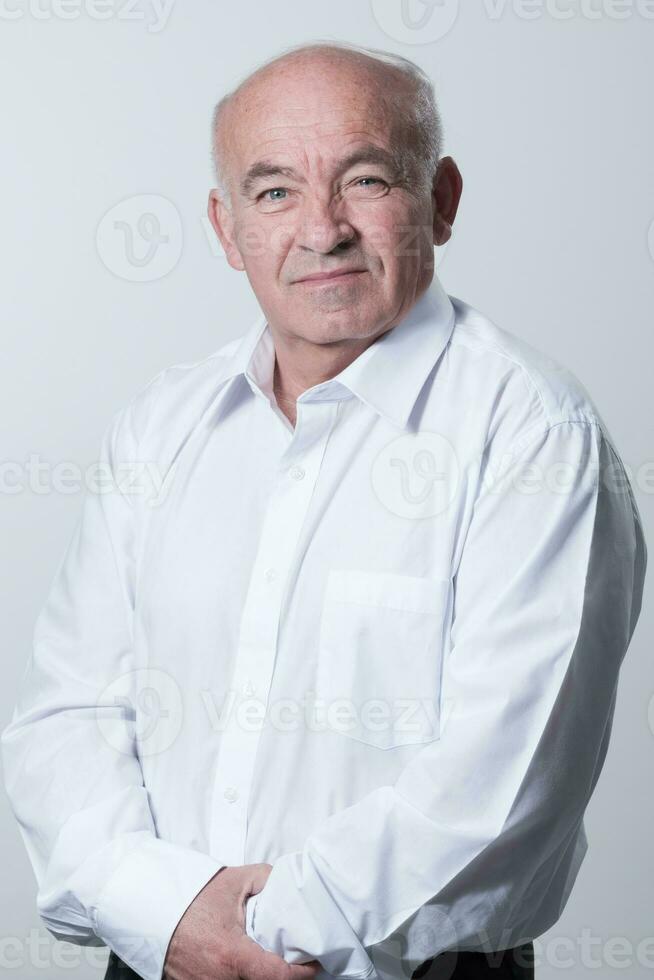 Portrait of an older man wearing a white shirt on a gray background. A healthy old man looks at a camera isolated over a gray wall. An older man smiles at the camera, a happy old man. photo