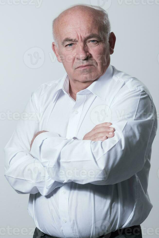 Confident senior man in white shirt crossing hands on chest and looking at camera while standing against gray background. Self confident senior isolated white studio shoot. photo