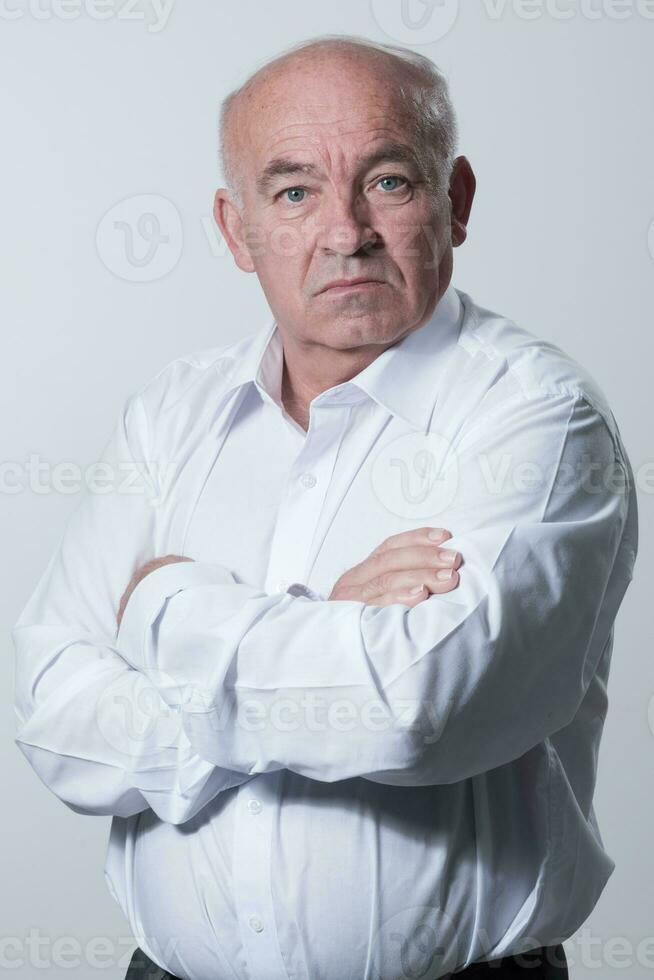 Confident senior man in white shirt crossing hands on chest and looking at camera while standing against gray background. Self confident senior isolated white studio shoot. photo