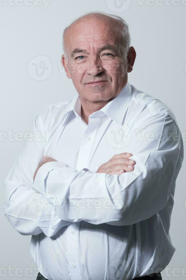 confidente mayor hombre en blanco camisa cruce manos en cofre y mirando a cámara mientras en pie en contra gris antecedentes. yo confidente mayor aislado blanco estudio disparo. foto