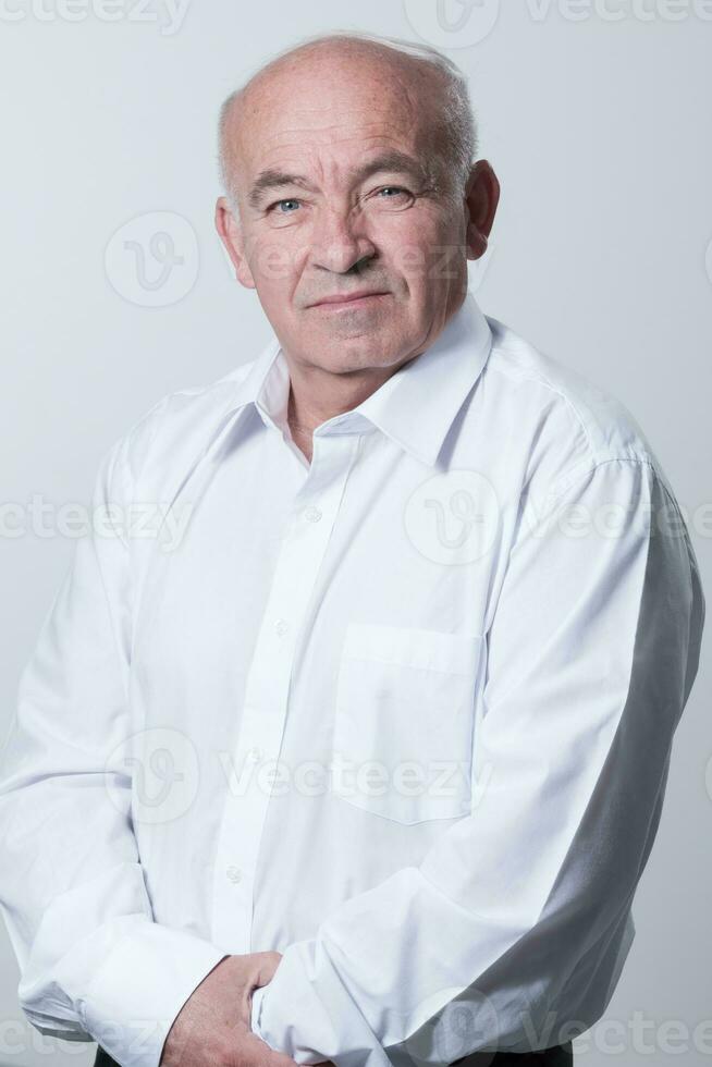 Portrait of an older man wearing a white shirt on a gray background. A healthy old man looks at a camera isolated over a gray wall. An older man smiles at the camera, a happy old man. photo