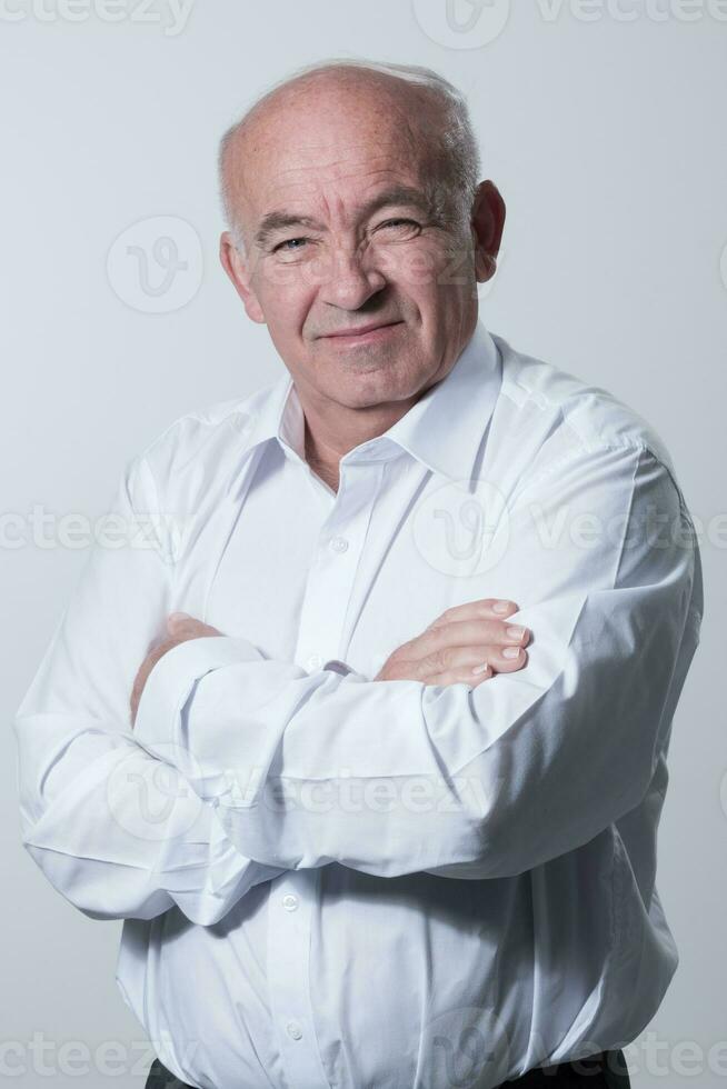 Confident senior man in white shirt crossing hands on chest and looking at camera while standing against gray background. Self confident senior isolated white studio shoot. photo