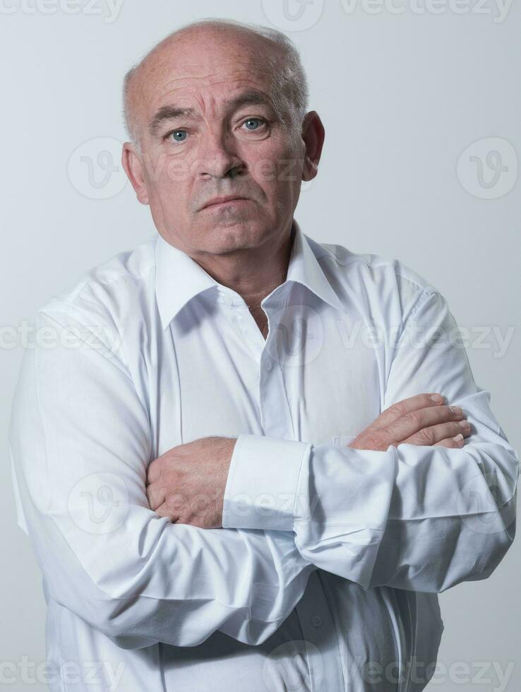 confidente mayor hombre en blanco camisa cruce manos en cofre y mirando a cámara mientras en pie en contra gris antecedentes. yo confidente mayor aislado blanco estudio disparo. foto