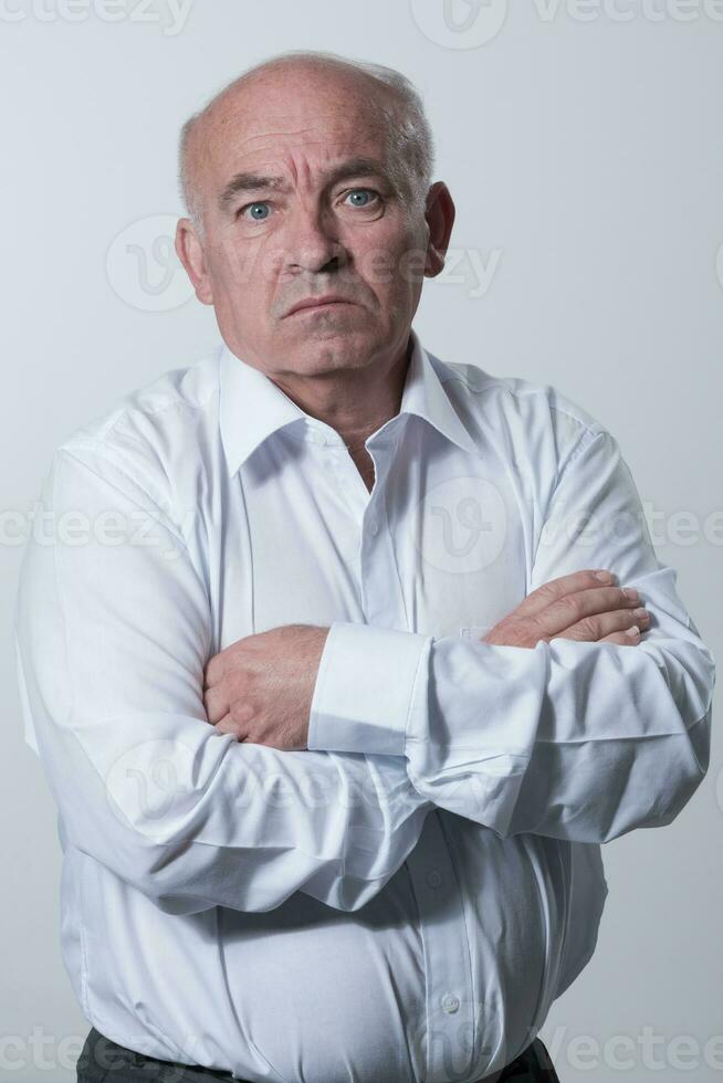 Confident senior man in white shirt crossing hands on chest and looking at camera while standing against gray background. Self confident senior isolated white studio shoot. photo