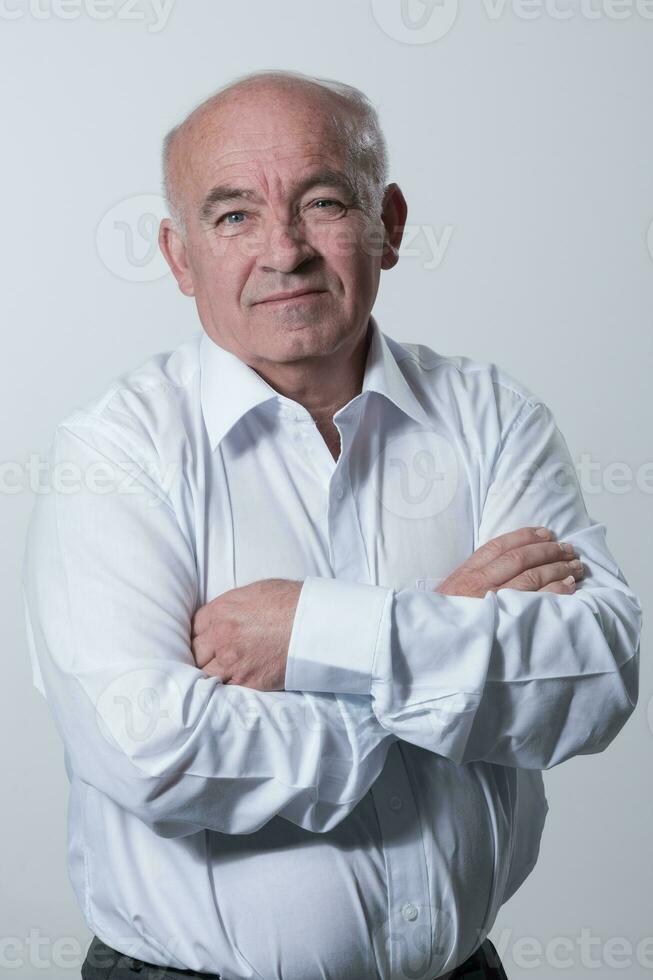 Confident senior man in white shirt crossing hands on chest and looking at camera while standing against gray background. Self confident senior isolated white studio shoot. photo