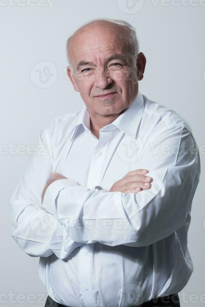 Confident senior man in white shirt crossing hands on chest and looking at camera while standing against gray background. Self confident senior isolated white studio shoot. photo