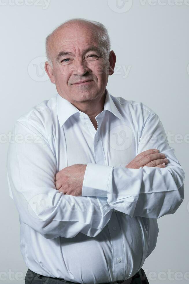 Confident senior man in white shirt crossing hands on chest and looking at camera while standing against gray background. Self confident senior isolated white studio shoot. photo