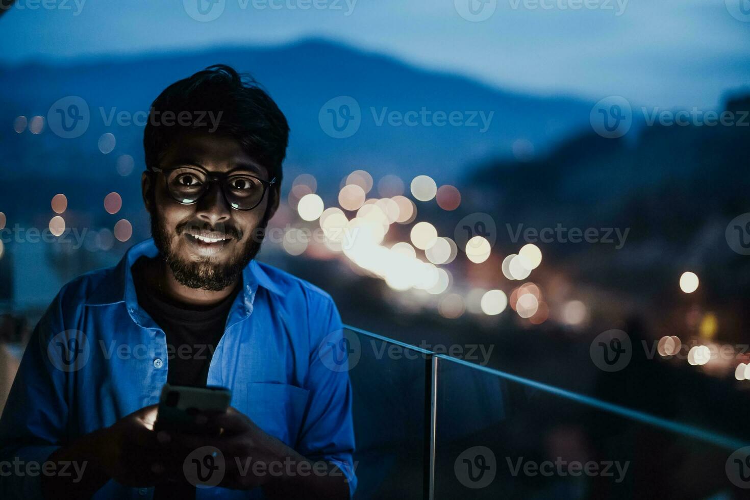 An indian man with glasses and a blue shirt using a smartphone at night on the balcony of a modern building. In the background of the night street of the city photo