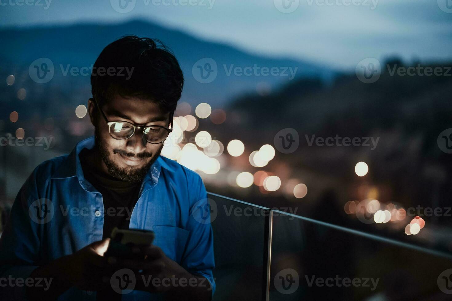 An indian man with glasses and a blue shirt using a smartphone at night on the balcony of a modern building. In the background of the night street of the city photo