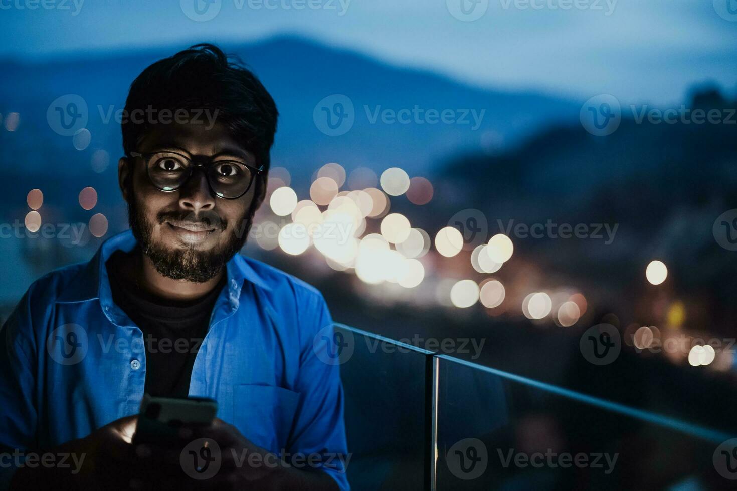An indian man with glasses and a blue shirt using a smartphone at night on the balcony of a modern building. In the background of the night street of the city photo