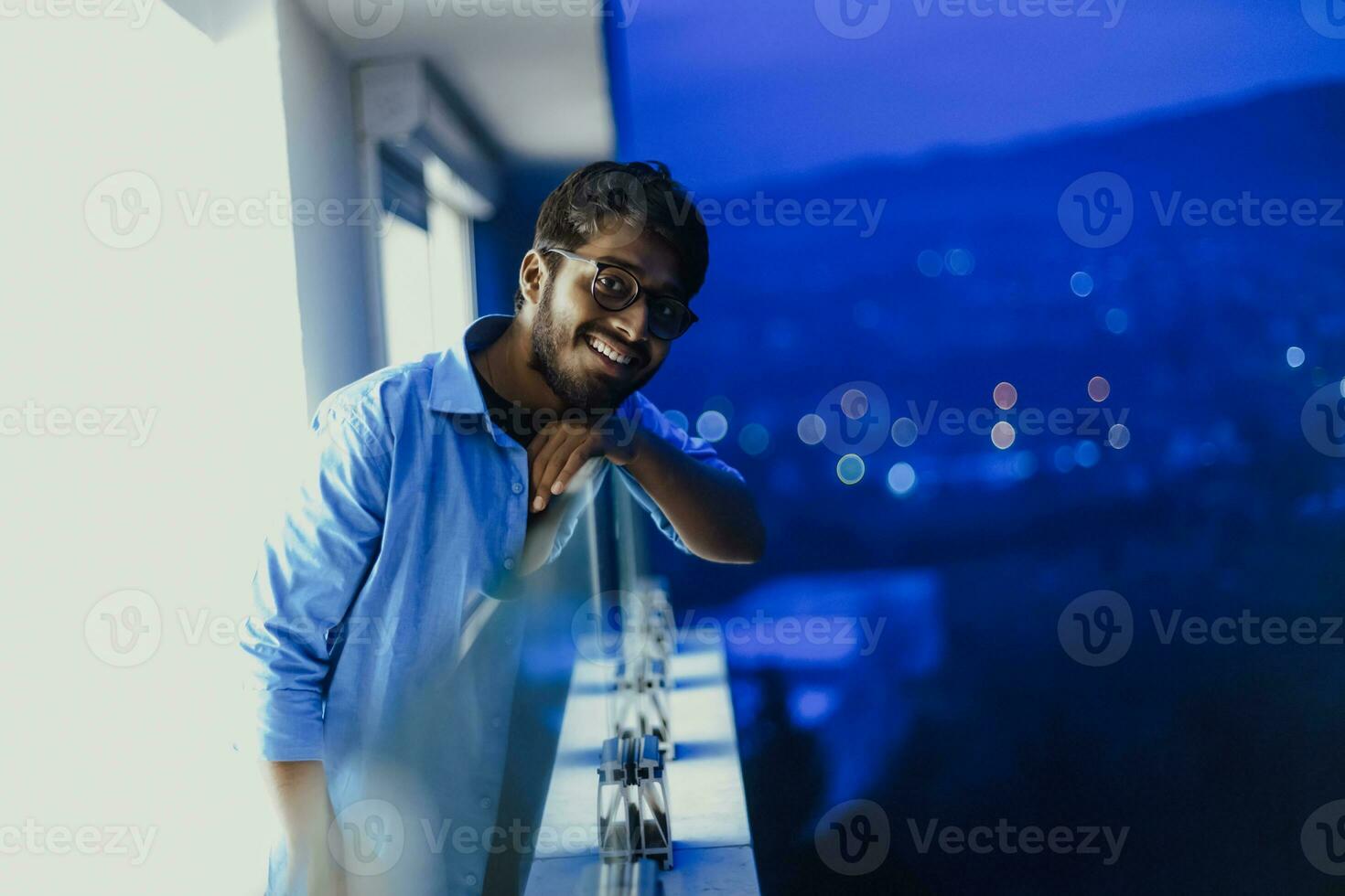 An Indian man with glasses and a blue shirt looks around the city at night. In the background of the night street of the city photo