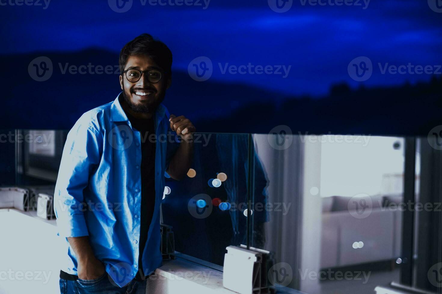 An Indian man with glasses and a blue shirt looks around the city at night. In the background of the night street of the city photo