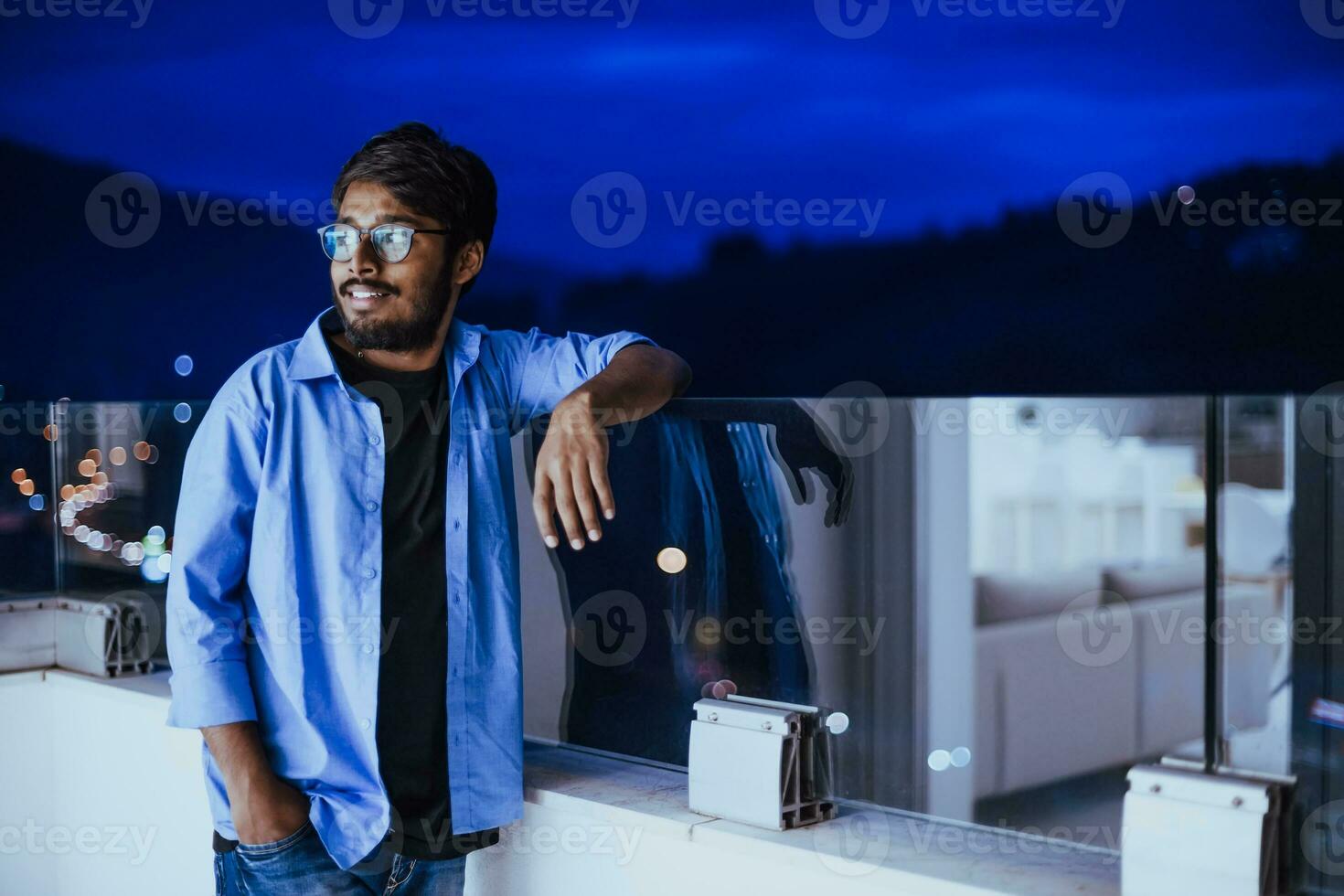 An Indian man with glasses and a blue shirt looks around the city at night. In the background of the night street of the city photo