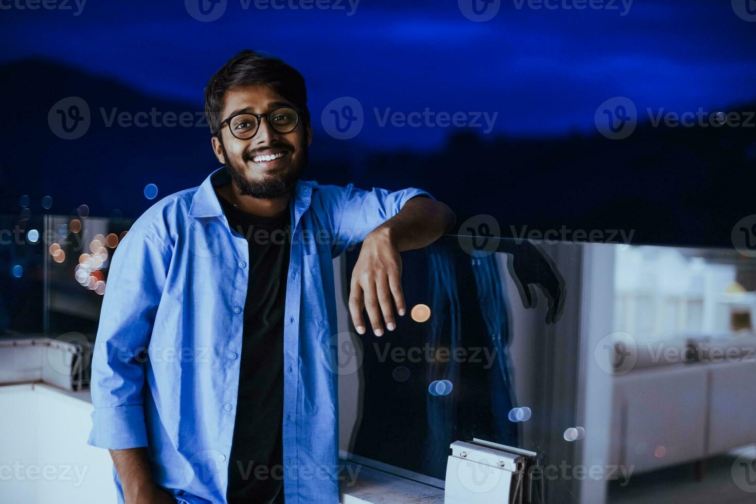 An Indian man with glasses and a blue shirt looks around the city at night. In the background of the night street of the city photo