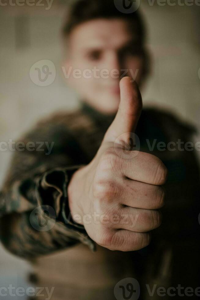 The soldier makes a gesture of success with his hand. A soldier in full war gear stands in front of a stone wall and shows the ok sign with his finger photo