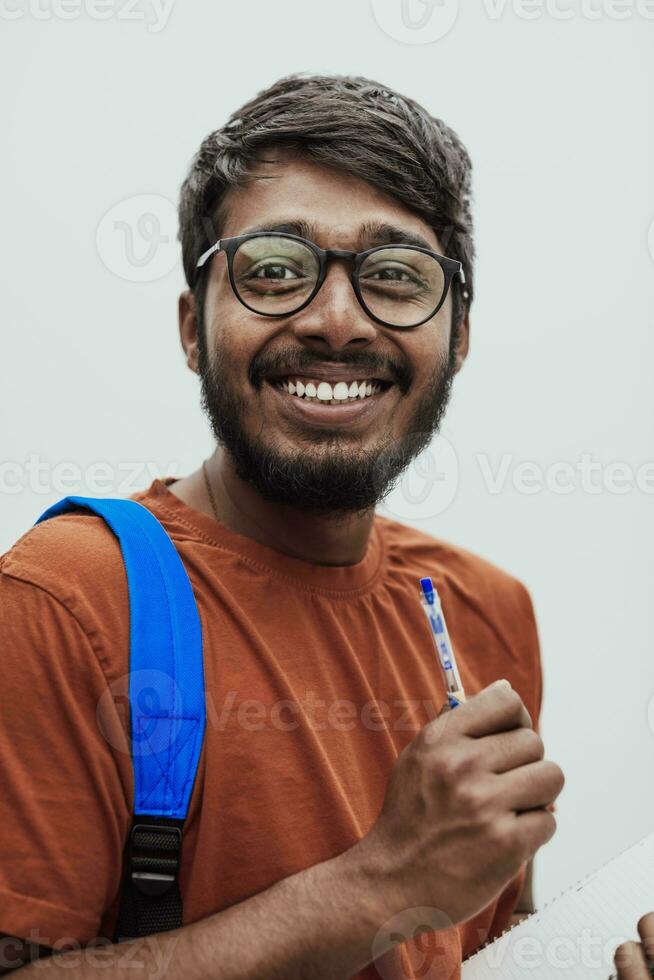 indio estudiante con azul mochila, lentes y cuaderno posando en gris antecedentes. el concepto de educación y enseñanza. hora a Vamos espalda a colegio foto