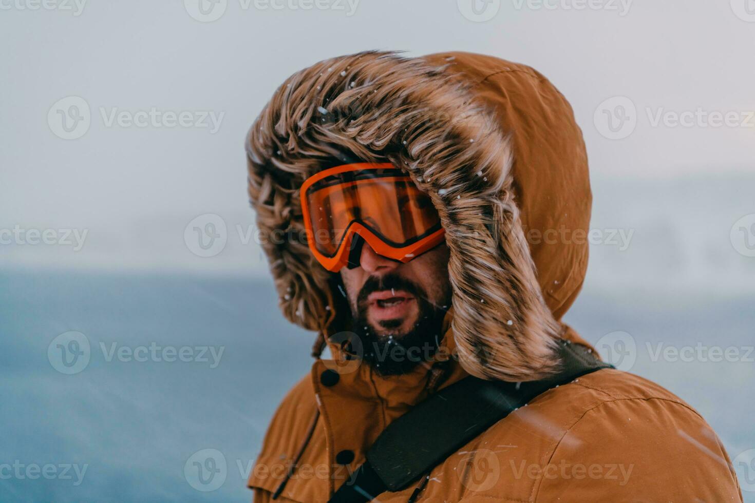 Headshot photo of a man in a cold snowy area wearing a thick brown winter jacket, snow goggles and gloves. Life in cold regions of the country.