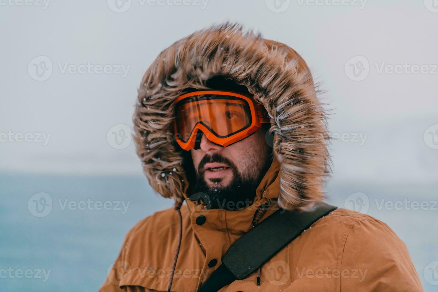 Headshot photo of a man in a cold snowy area wearing a thick brown winter jacket, snow goggles and gloves. Life in cold regions of the country.