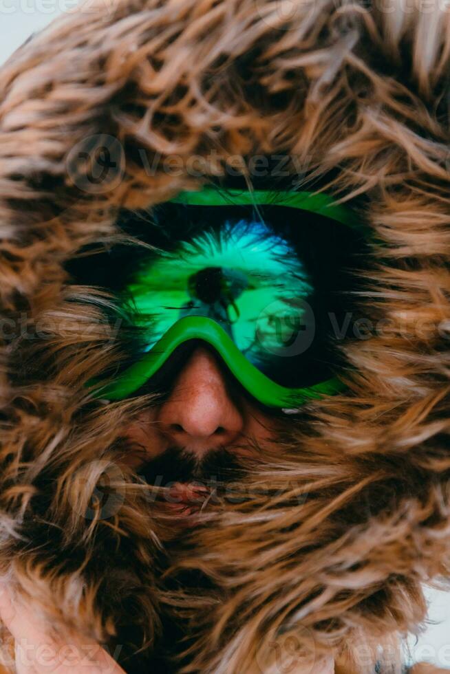 disparo a la cabeza foto de un hombre en un frío Nevado zona vistiendo un grueso marrón invierno chaqueta, nieve gafas de protección y guantes. vida en frío regiones de el país.