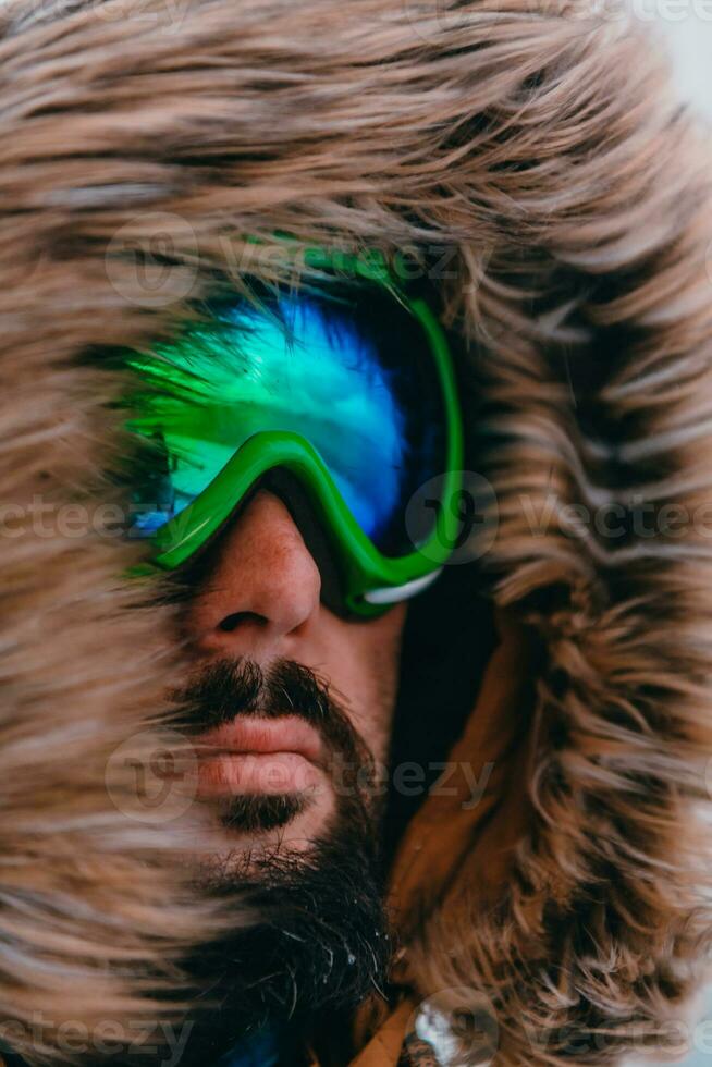Headshot photo of a man in a cold snowy area wearing a thick brown winter jacket, snow goggles and gloves. Life in cold regions of the country.
