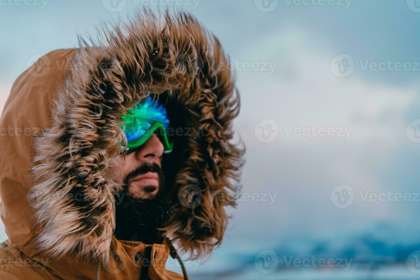 Headshot photo of a man in a cold snowy area wearing a thick brown winter jacket, snow goggles and gloves. Life in cold regions of the country.
