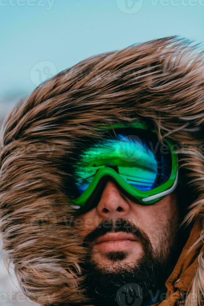 Headshot photo of a man in a cold snowy area wearing a thick brown winter jacket, snow goggles and gloves. Life in cold regions of the country.