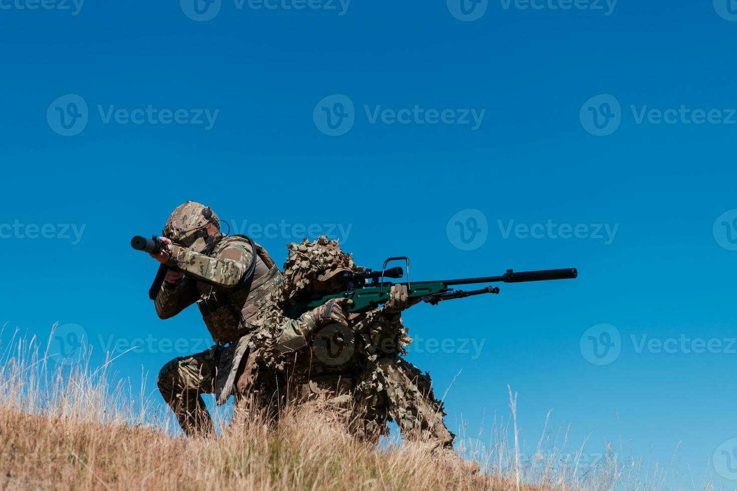 A sniper team squad of soldiers is going undercover. Sniper assistant and team leader walking and aiming in nature with yellow grass and blue sky. Tactical camouflage uniform. photo
