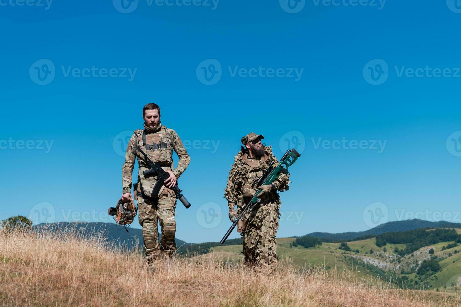 A sniper team squad of soldiers is going undercover. Sniper assistant and team leader walking and aiming in nature with yellow grass and blue sky. Tactical camouflage uniform. photo