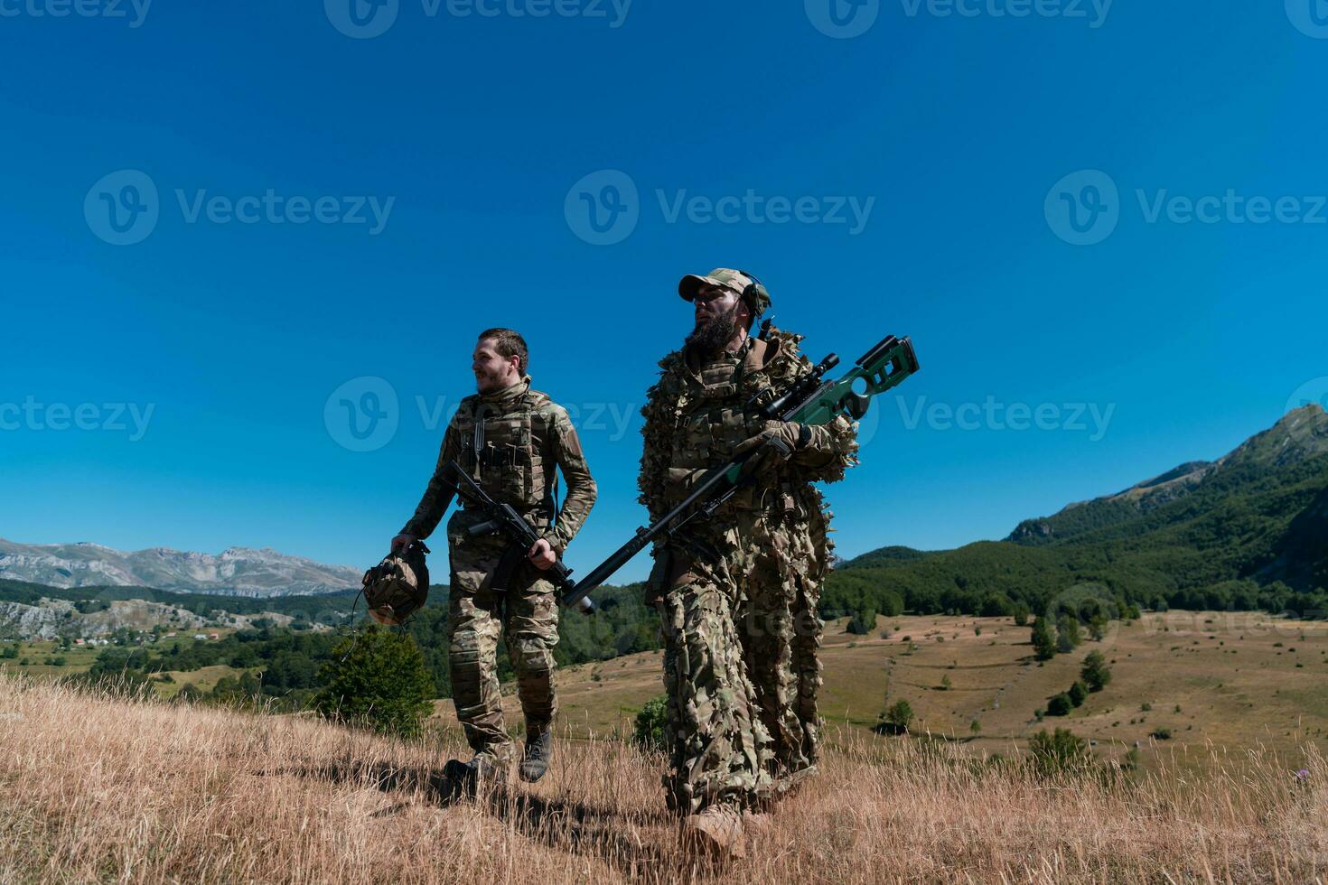 A sniper team squad of soldiers is going undercover. Sniper assistant and team leader walking and aiming in nature with yellow grass and blue sky. Tactical camouflage uniform. photo