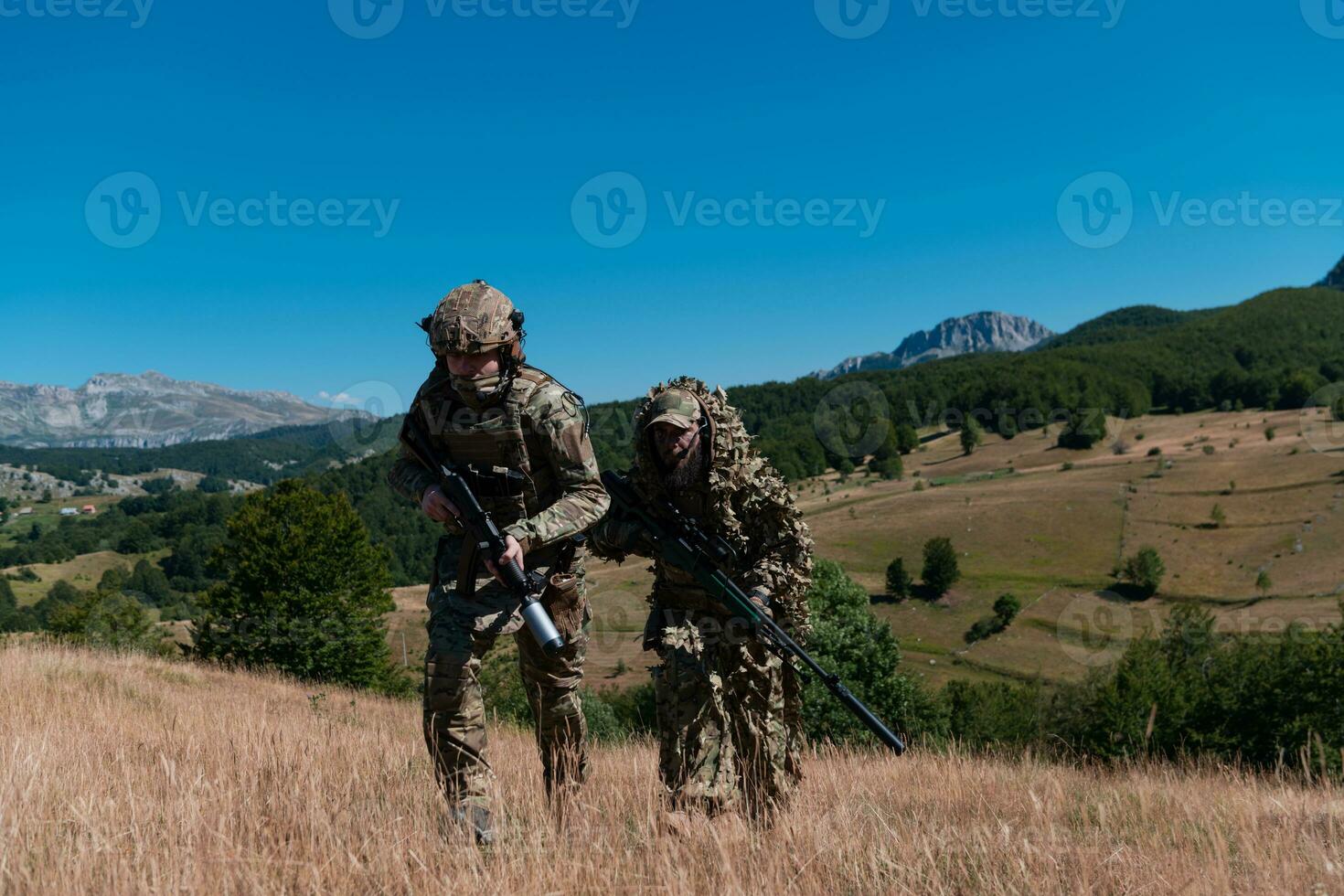 A sniper team squad of soldiers is going undercover. Sniper assistant and team leader walking and aiming in nature with yellow grass and blue sky. Tactical camouflage uniform. photo