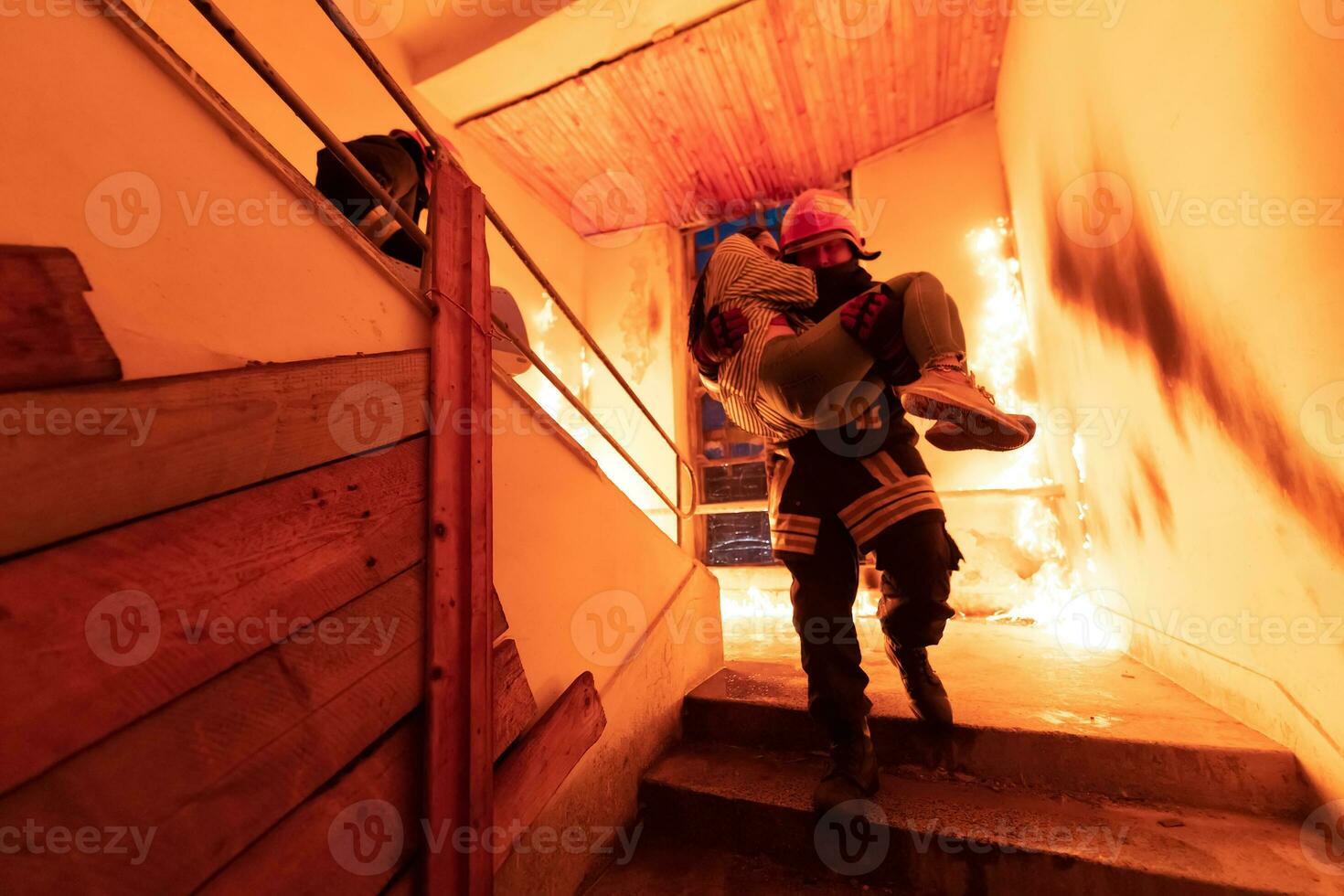 Brave Fireman Descends Stairs of a Burning Building and Holds Saved Girl in His Arms. Open fire and one Firefighter in the Background. photo