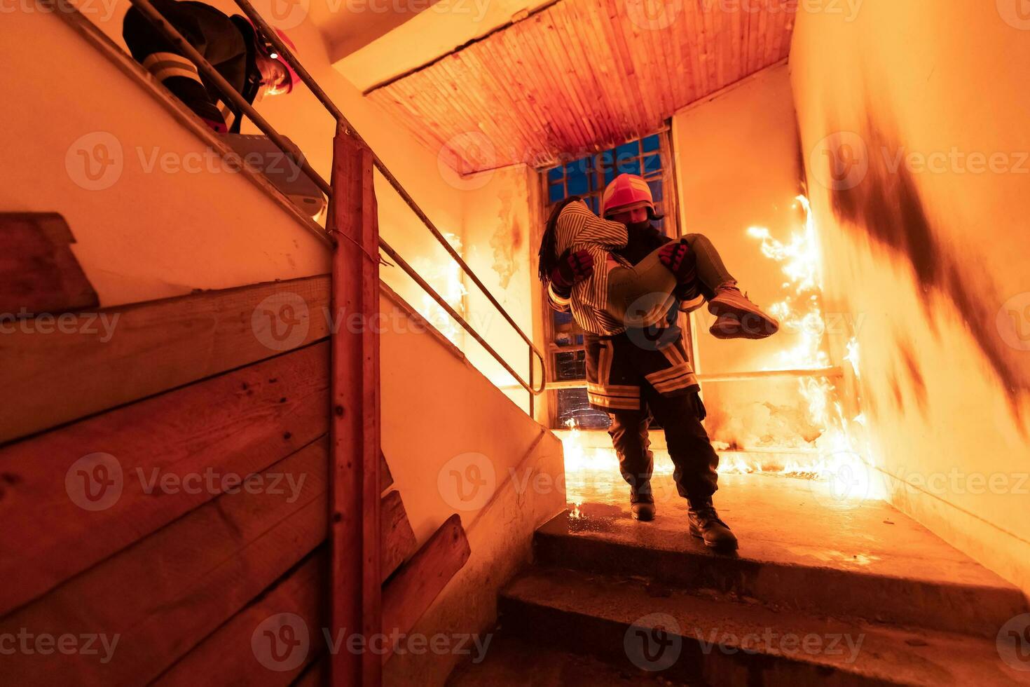 Brave Fireman Descends Stairs of a Burning Building and Holds Saved Girl in His Arms. Open fire and one Firefighter in the Background. photo