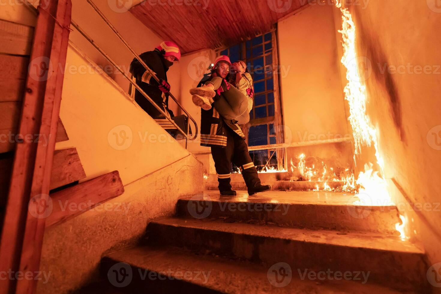 Brave Fireman Descends Stairs of a Burning Building and Holds Saved Girl in His Arms. Open fire and one Firefighter in the Background. photo