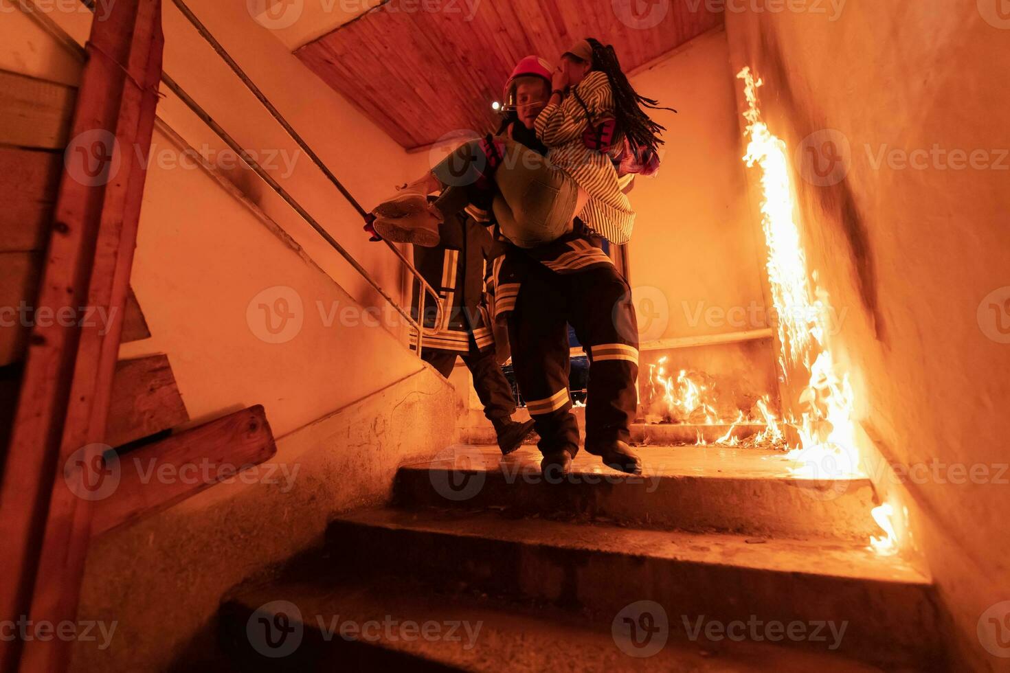 Brave Fireman Descends Stairs of a Burning Building and Holds Saved Girl in His Arms. Open fire and one Firefighter in the Background. photo