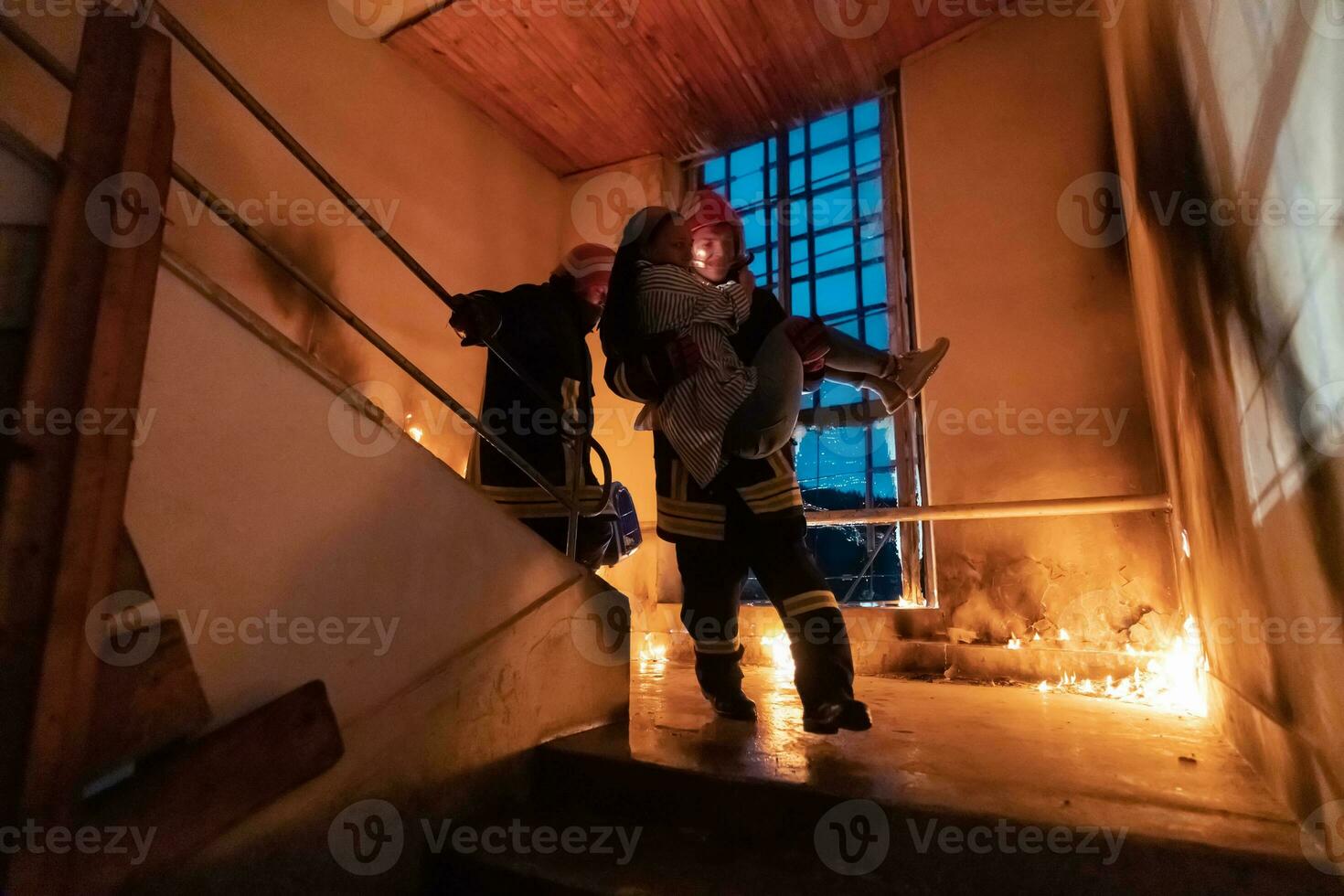 valiente bombero desciende escalera de un ardiente edificio y sostiene salvado niña en su brazos. abierto fuego y uno bombero en el antecedentes. foto