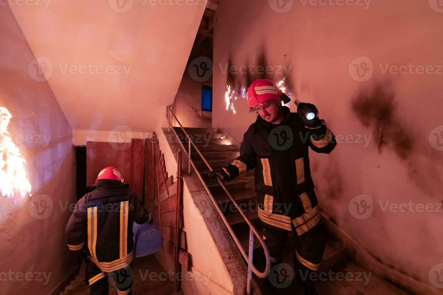 Brave Fireman Descends Stairs of a Burning Building and Holds Saved Girl in His Arms. Open fire and one Firefighter in the Background. photo