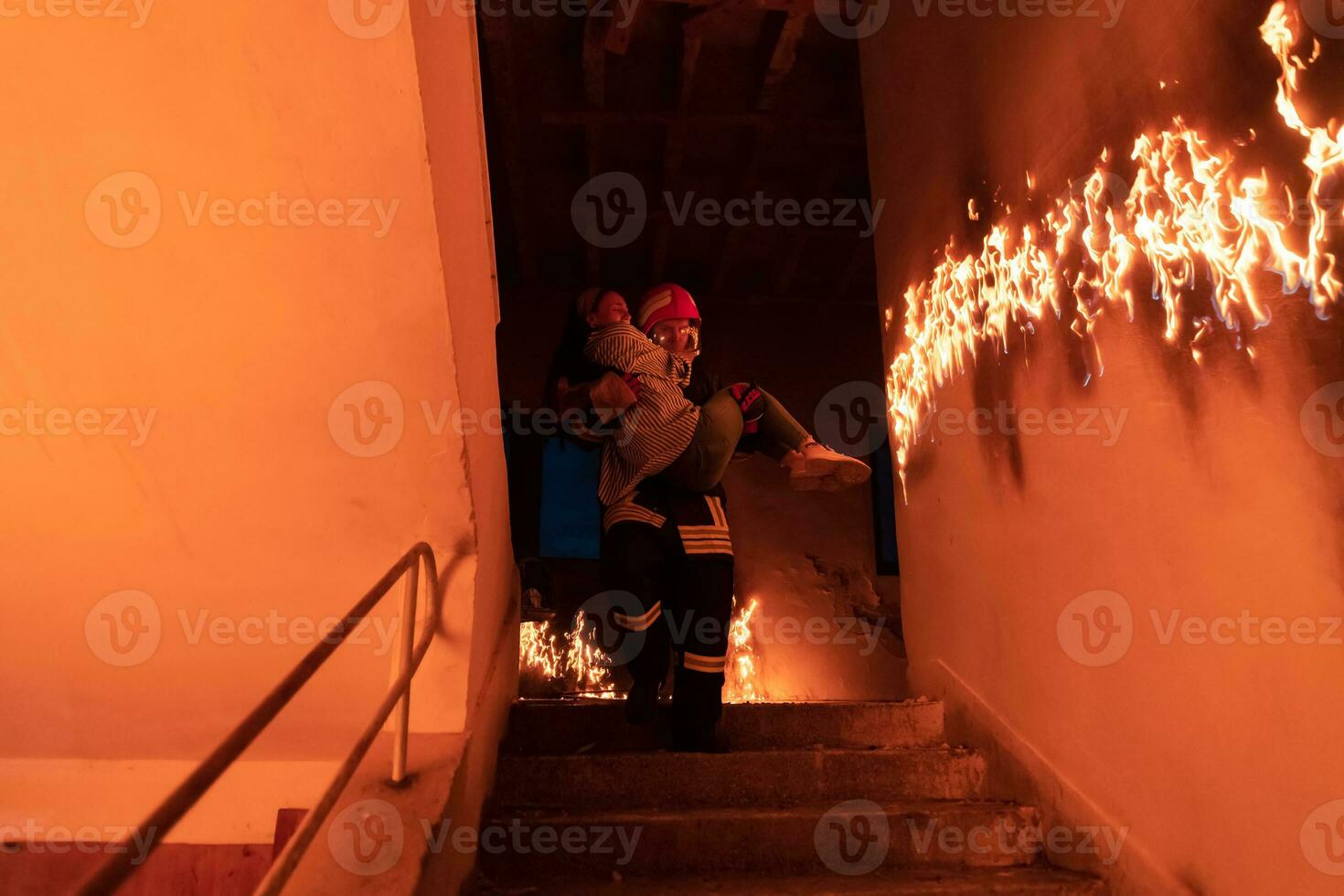 Brave Fireman Descends Stairs of a Burning Building and Holds Saved Girl in His Arms. Open fire and one Firefighter in the Background. photo