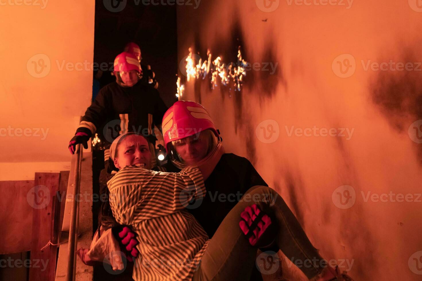 Brave Fireman Descends Stairs of a Burning Building and Holds Saved Girl in His Arms. Open fire and one Firefighter in the Background. photo