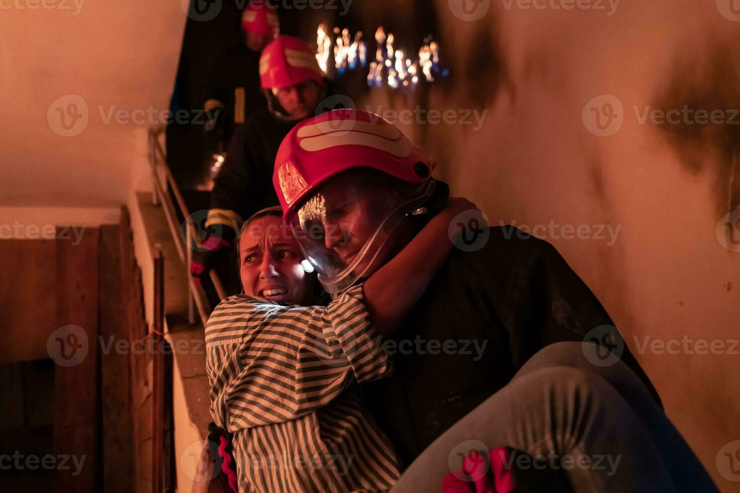 Brave Fireman Descends Stairs of a Burning Building and Holds Saved Girl in His Arms. Open fire and one Firefighter in the Background. photo